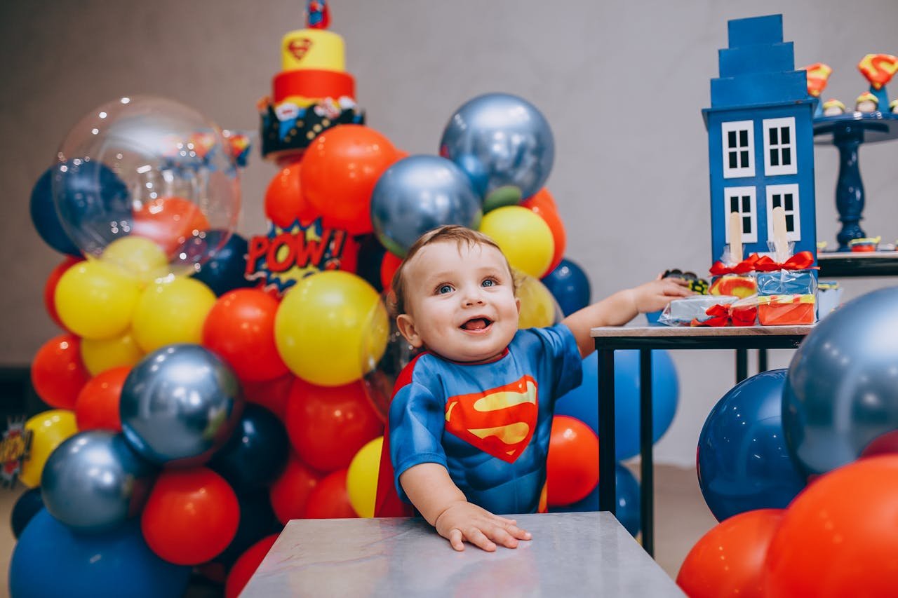 Adorable baby in superhero costume celebrating a colorful birthday surrounded by balloons and cake.