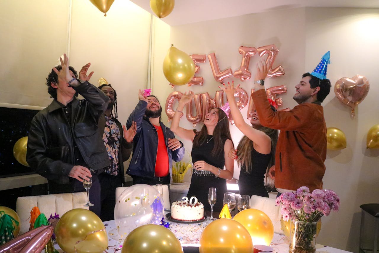 brand-01 Group of friends celebrating a birthday indoors, surrounded by balloons and cake.